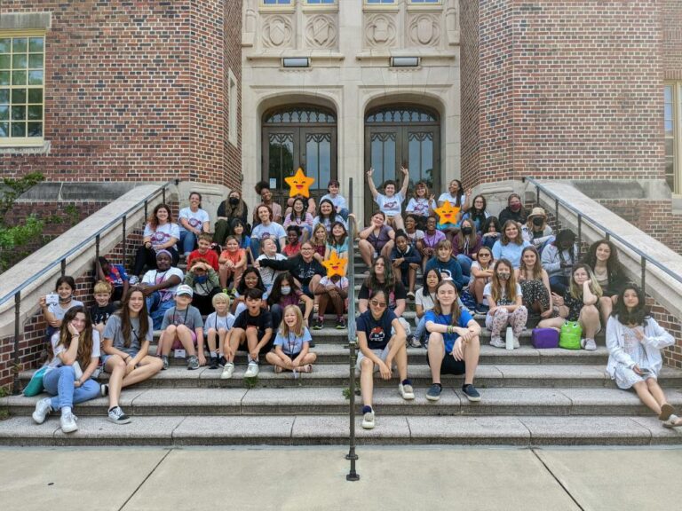 A group of school aged children pose on the steps of a large building.