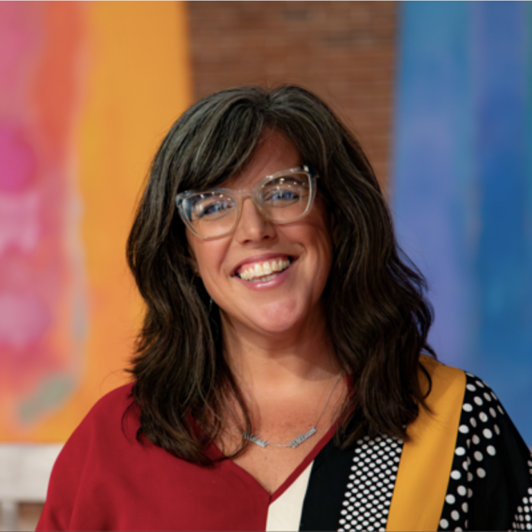 a smiling woman with glasses standing in front of colorful paintings